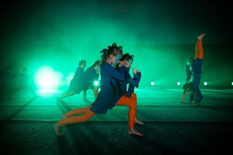 Tanzende Kinder in blauen und orangefarbenen Outfits auf einer beleuchteten B&uuml;hne mit gr&uuml;nem Licht und Nebel.