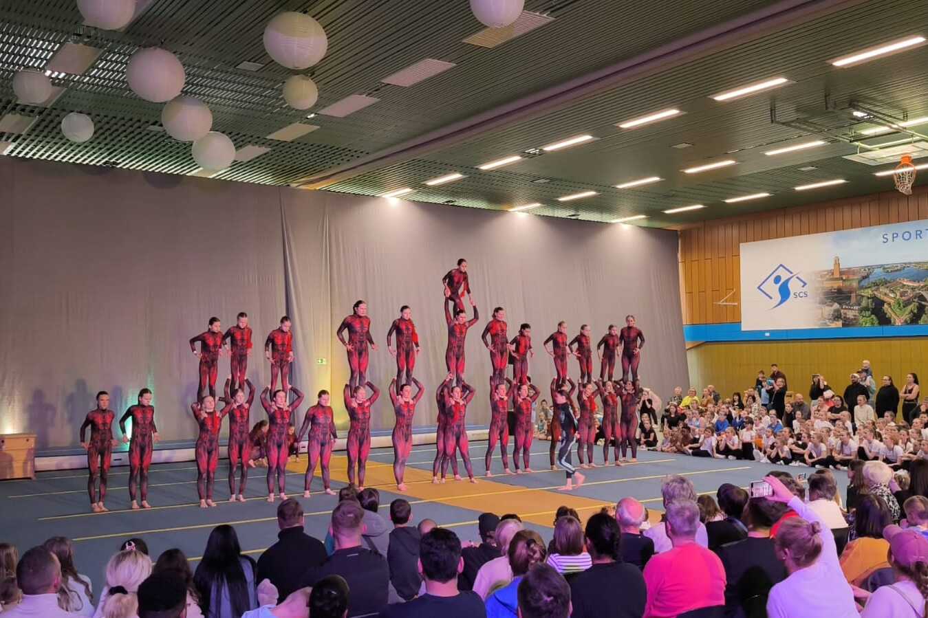 Akrobatikaufführung mit mehreren Artisten in Rot und Schwarz, die pyramidenförmig auf einer Matte stehen, Zuschauer im Hintergrund.