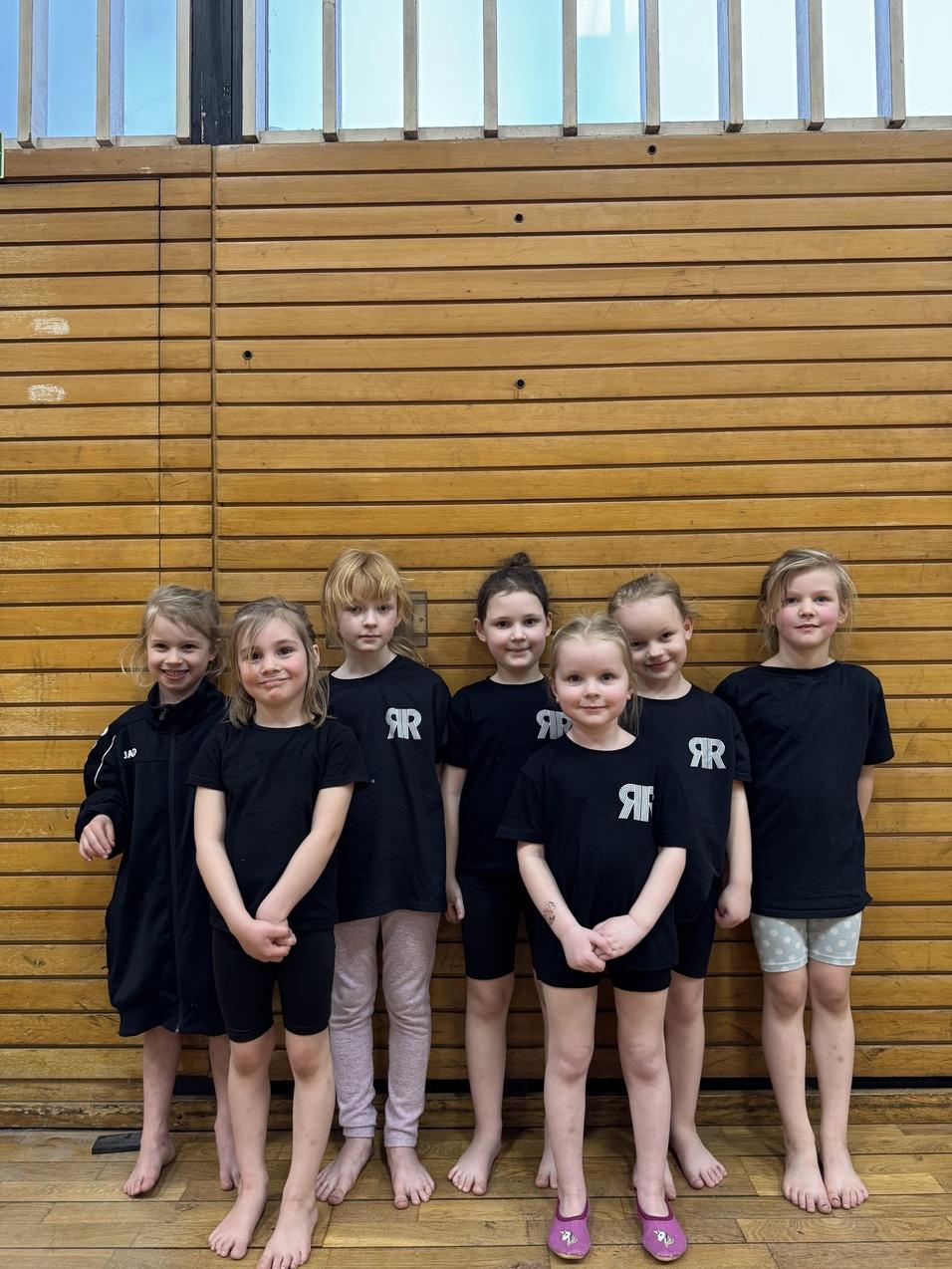 Gruppe von sieben Mädchen in schwarzen T-Shirts und Sporthosen vor einer Holzwand in einer Turnhalle.