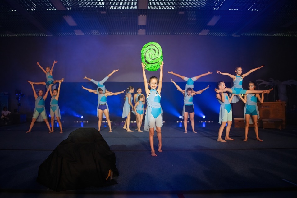Acht weibliche Akrobatinnen in blauen Kostümen führen auf einer Bühne mit Lichtern eine Gruppenübung durch.