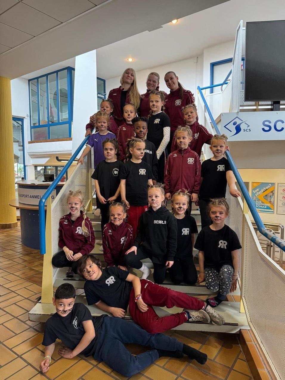 Gruppe von Kinderturnern in schwarzen und roten Outfits mit Gesichtern in Tiermotiv auf einer Treppe.