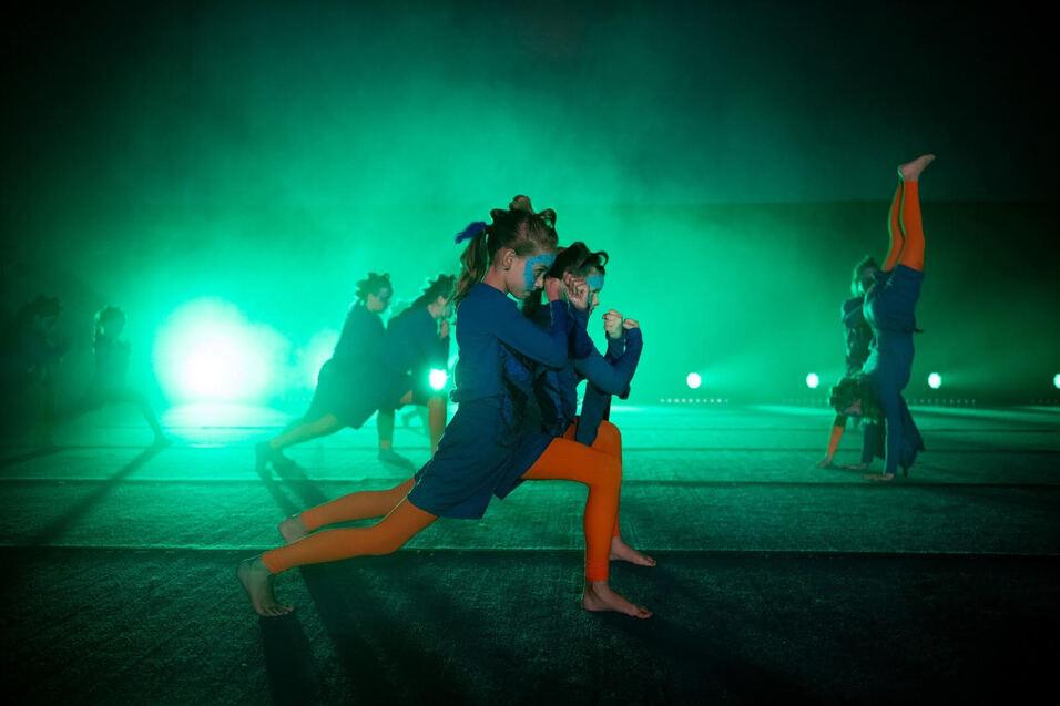Gruppe von T&auml;nzerinnen in blauen Outfits und orangen Leggings pr&auml;sentiert synchronisierte Bewegungen auf einer B&uuml;hne.