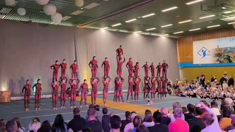 Akrobatikaufführung mit mehreren Artisten in Rot und Schwarz, die pyramidenförmig auf einer Matte stehen, Zuschauer im Hintergrund.