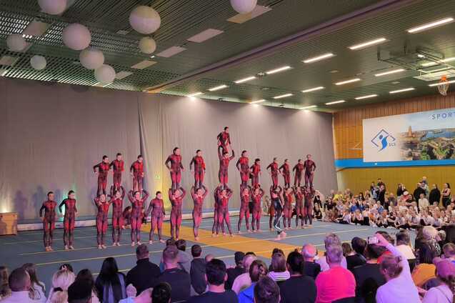 Akrobatikaufführung mit mehreren Artisten in Rot und Schwarz, die pyramidenförmig auf einer Matte stehen, Zuschauer im Hintergrund.