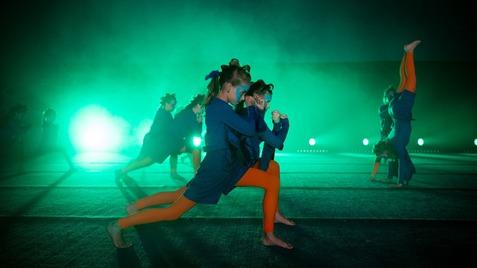 Tanzende Kinder in blauen und orangefarbenen Outfits auf einer beleuchteten Bühne mit grünem Licht und Nebel.