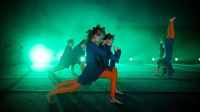 Tanzende Kinder in blauen und orangefarbenen Outfits auf einer beleuchteten Bühne mit grünem Licht und Nebel.