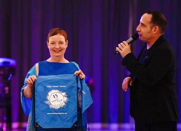 Eine Frau in blauer Kleidung hält ein Geschenk mit dem Logo eines Tango-Seminars, während ein Mann im Anzug spricht.