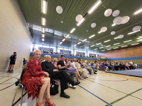 Blick auf eine Menschenmenge in einem Sporthall, vor allem ältere Zuschauer sitzen auf Stühlen, bedeckt mit bunten Dekorationen.