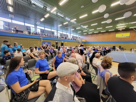Voll besetzte Halle mit Zuschauern in blauen T-Shirts, die auf einer Veranstaltung im Sitzen warten.