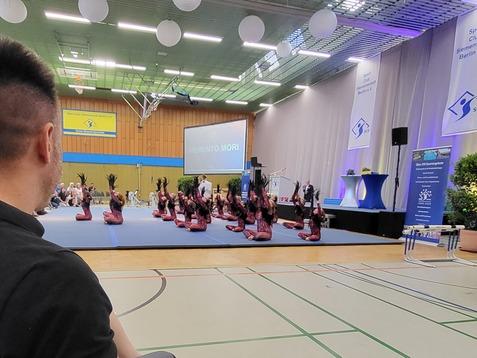 Blick auf eine Sportveranstaltung mit Menschen auf Stühlen und einer Aufführung im Hintergrund in einer Halle.