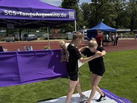 Zwei Mädchen tanzen auf einem großen, beschrifteten Tanzmatte vor einem lila Zelt mit dem Logo von SCS Tango Argentino.