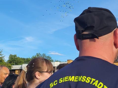 Bunte Luftballons steigen vor einem blauen Himmel über einer Menschenmenge auf, sichtbar sind Rücken von Teilnehmern.
