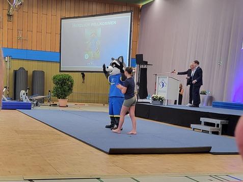 Eine Person in einem Tierkostüm steht auf einer Bühne, während ein Redner an einem Pult spricht. Zuschauer im Vordergrund.