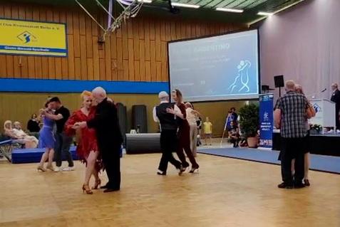 Tanzende Paare in eleganter Kleidung bei einer Veranstaltung in einer Sporthalle, im Hintergrund eine Präsentationsleinwand.