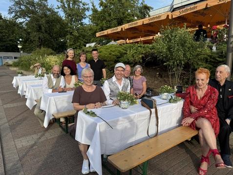 Gruppenszene mit neun Personen an einer gedeckten Tafel draußen, umgeben von Pflanzen und Sonnenschirmen.