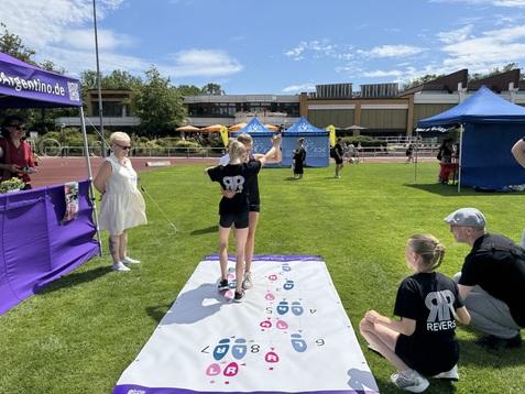 Zwei Kinder stehen auf einer Spielfläche mit Symbolen, während Erwachsene in sportlicher Umgebung zuschauen.