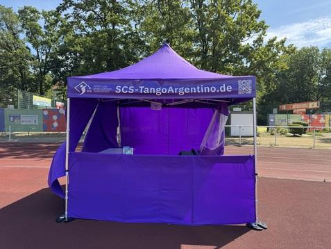 Lila Infostand des SCS-TangoArgentino auf einem Sportplatz mit Bäumen im Hintergrund.