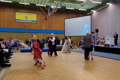 Tanzende Paare in festlicher Kleidung auf einer Bühne, Zuschauer sitzen an der Seite und beobachten die Darbietung.