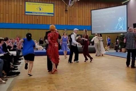 Tanzende Paare auf einer Bühne, umgeben von applaudierendem Publikum in einem Veranstaltungsraum.