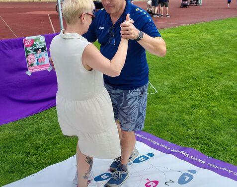 Tanzende Personen stehen auf einer farbigen Matte mit Fuß- und Zahlenmustern im Freien bei einer Veranstaltung.
