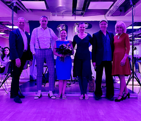 Sechs Personen stehen zusammen auf einer Bühne, eine Frau hält einen Blumenstrauß, fröhliche Stimmung im Hintergrund.