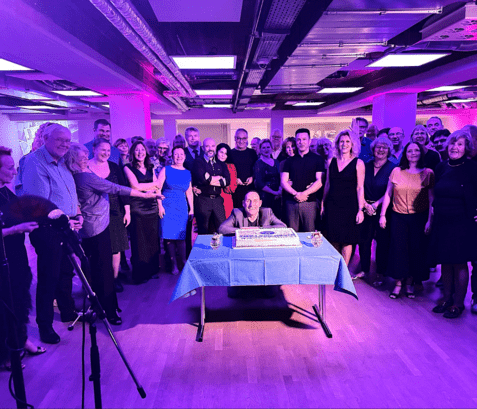 Gruppenfoto von Gästen bei einer Feier, um einen Tisch mit einer Torte und blauem Tuch versammelt.