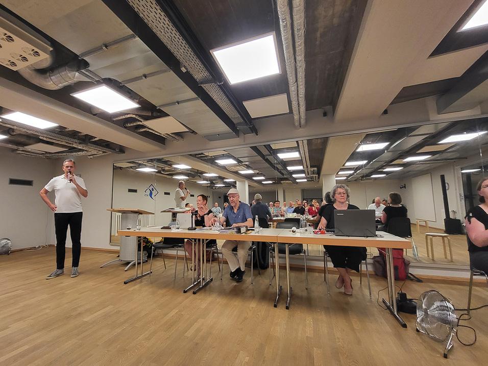 Eine Person steht mit Mikrofon, während am Tisch mehrere Menschen sitzen und auf eine Bildschirmpräsentation blicken.