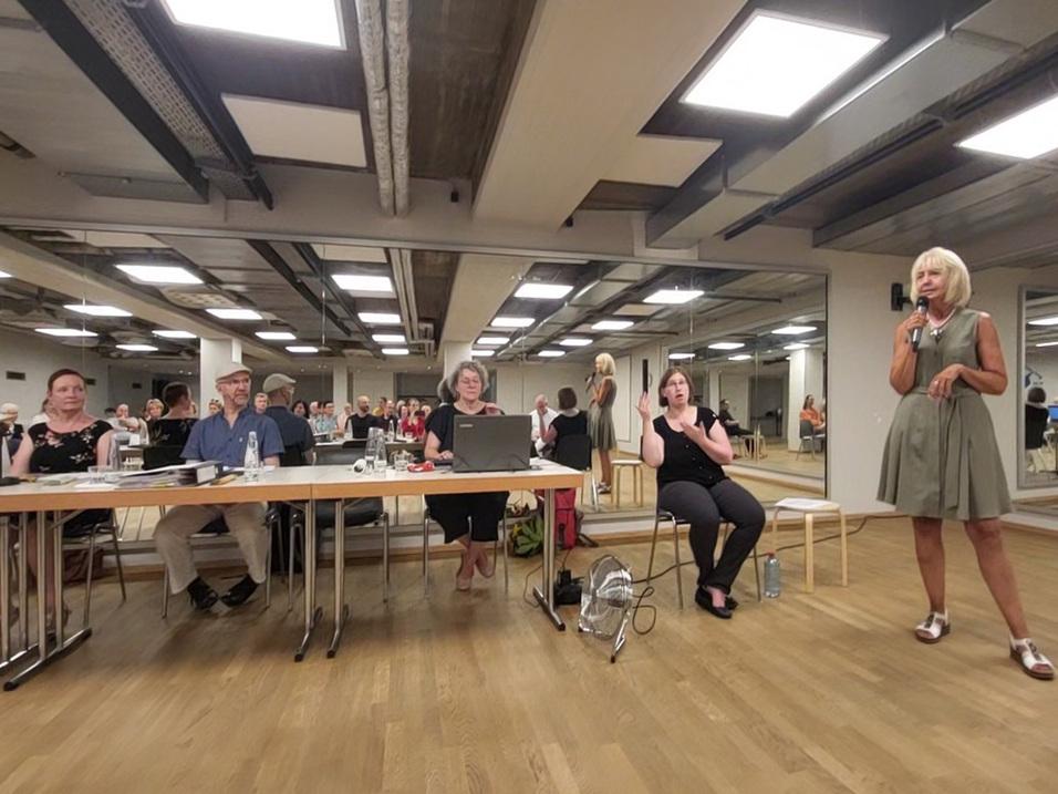 Frauen und Männer sitzen an Tischen in einem Raum, während eine Frau im Vordergrund mit Mikrofon spricht.