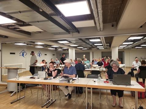 Versammlung in einem modernen Raum mit mehreren Tischen, Teilnehmern und einem Rednerpult im Fokus.