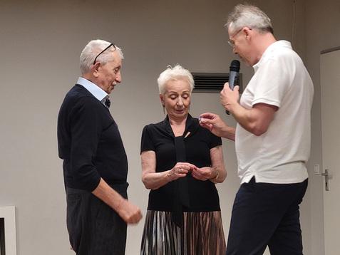 Drei Personen stehen auf einer Bühne, während einer mit einem Mikrofon spricht und ein Objekt zeigt.