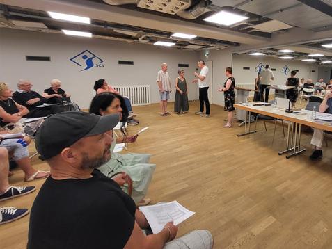 Menschen in einem Raum stehen und sitzen bei einer Veranstaltung, einige halten Blätter, Tische im Hintergrund.