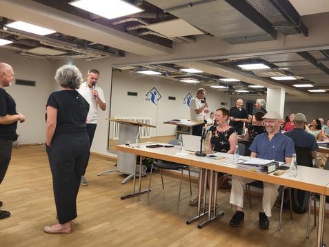 Gruppenszene bei einer Veranstaltung, mit zwei Personen am Mikrofon und Publikum im Hintergrund. Tische und Notebooks sichtbar.