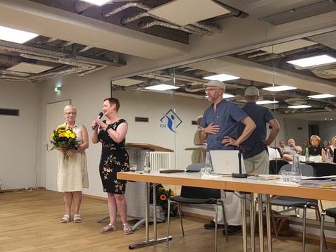 Zwei Frauen stehen auf einer Bühne; eine hält einen Blumenstrauß, während die andere spricht. Männer und Frauen im Publikum.