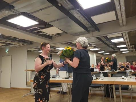 Zwei Frauen stehen sich gegenüber, eine hält einen Blumenstrauß, während die andere ein Mikrofon hält. Publikum im Hintergrund.