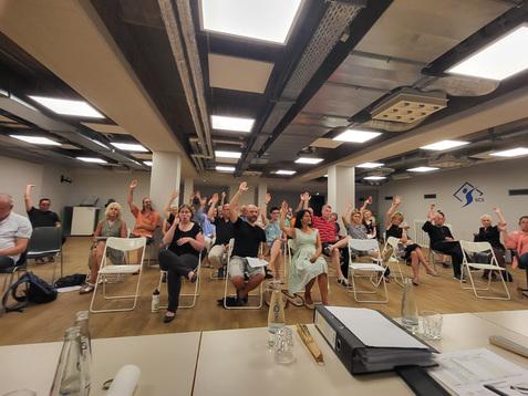 Mehrere Personen in einem Sitzungsraum heben ihre Hände zur Abstimmung. Tische und Stühle sind sichtbar.