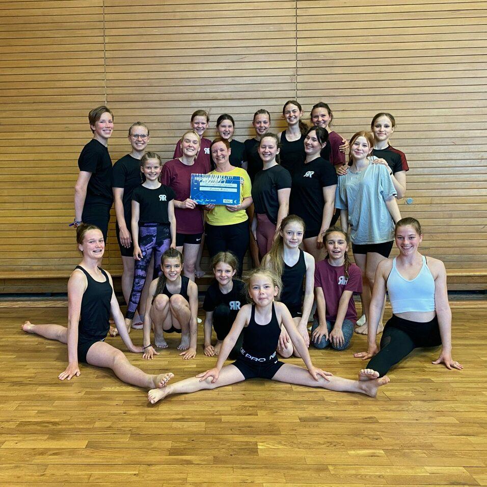 Gruppe von 22 Turnerinnen in Sportkleidung posieren lächelnd in einer Turnhalle, einige im Spagat und mit einer Auszeichnung.