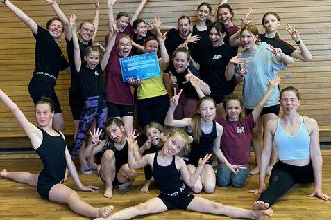 Gruppenselfie von 20 Tänzerinnen in Sportkleidung, die fröhlich posieren und eine Auszeichnung halten. Holzwand im Hintergrund.