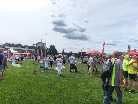 Menschen in bunten T-Shirts und Hüten auf einer Wiese, umgeben von Ständen und Zelten bei einer Veranstaltung.