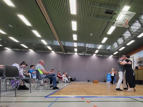 Tanzende Paare auf einer Holzfläche in einer Halle, Zuschauer sitzen auf Stühlen im Hintergrund.