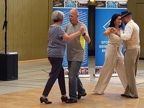 Zwei Paare tanzen in einer Sporthalle, im Hintergrund Banner mit Informationen und eine Lautsprecherbox.