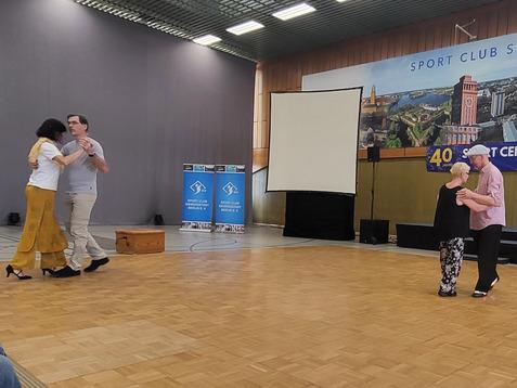 Tanzende Paare in einem Sportzentrum, im Hintergrund Banner des Sport Clubs und eine große Leinwand sichtbar.