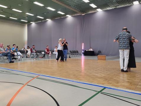 Tanzpaar auf einer Bühne mit Holzfußboden, Publikum sitzt auf Stühlen im Hintergrund, graue Wand im Hintergrund.