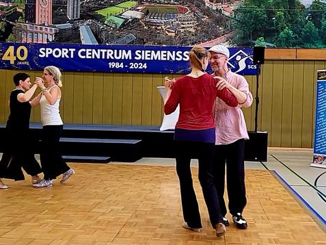 Tanzende Paare in einem Sportzentrum; Hintergrund zeigt eine Präsentation mit einem Stadtbild.