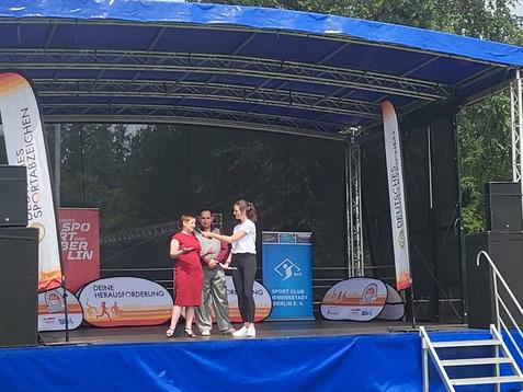 Drei Personen stehen auf einer Bühne, während eine Frau in rotem Kleid spricht und einen Mikrofon hält.