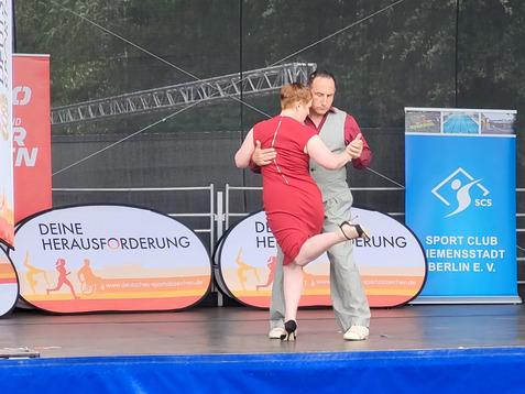 Tanzendes Paar auf einer Bühne, umgeben von Werbebannern, im Grüntönen des Hintergrunds.