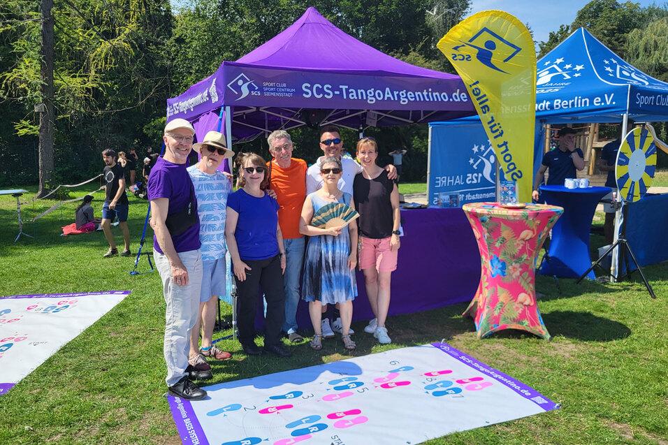 Gruppe von sechs Menschen mit bunten T-Shirts vor einem Zelt, umgeben von bunten Bannern und Spielstationen im Freien.