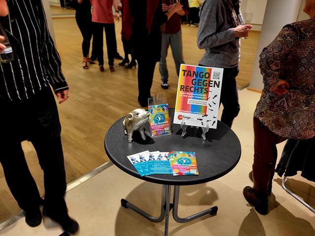 Tisch mit einem Flyer für „Tango gegen Rechts“ und einer kleinen Statue, umgeben von tanzenden Menschen im Hintergrund.