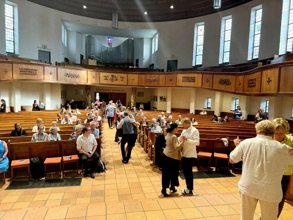 Vielzahl von Personen in einer Kirche, interagierend und sich bewegend, mit Holzbankreihen im Vordergrund.