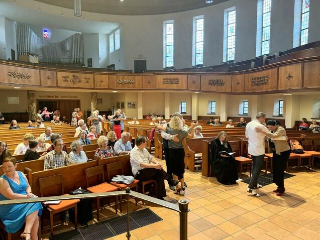 Tanzende Paare in einer Kirche, Zuschauer sitzen auf Holzbankreihen, farbiges Licht durch Buntglasfenster.