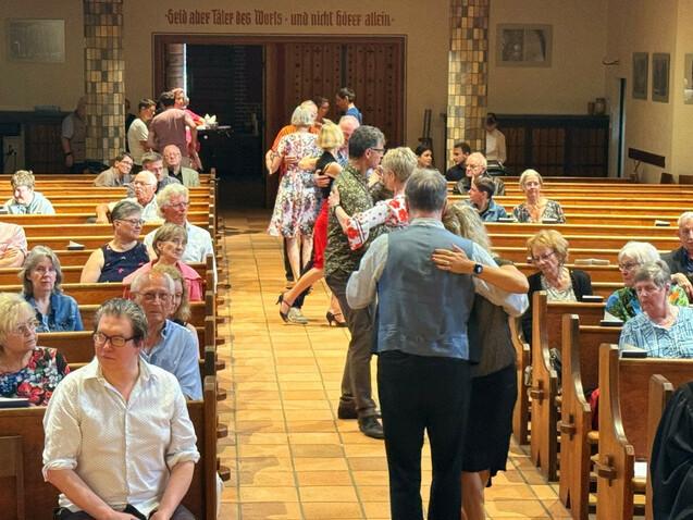 Tanzende Paare in einer Kirche umgeben von Sitzplätzen voller Zuschauer, die das Geschehen beobachten.